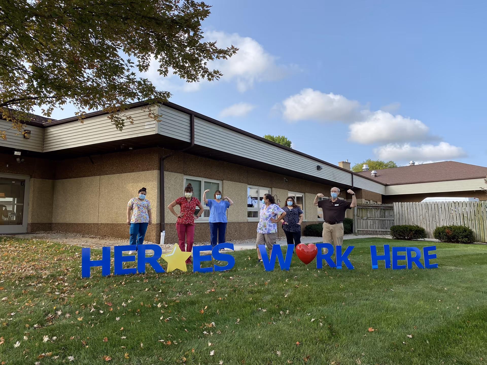 Six healthcare workers wearing masks stand outside a building on a grassy lawn behind large blue letters that spell out 'HEROES WORK HERE' with a yellow star replacing the letter O in HEROES and a red heart replacing the letter O in WORK. The sky is partly cloudy.