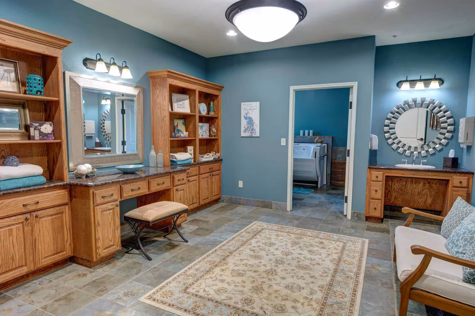 A spacious room with teal walls and tiled floor featuring wooden cabinetry with granite countertops, a cushioned bench, a decorative rug, a round mirror with a sunburst frame, and a white cushioned chair with patterned pillows. Through an open doorway, a bathroom with a walk-in bathtub and candles is visible.
