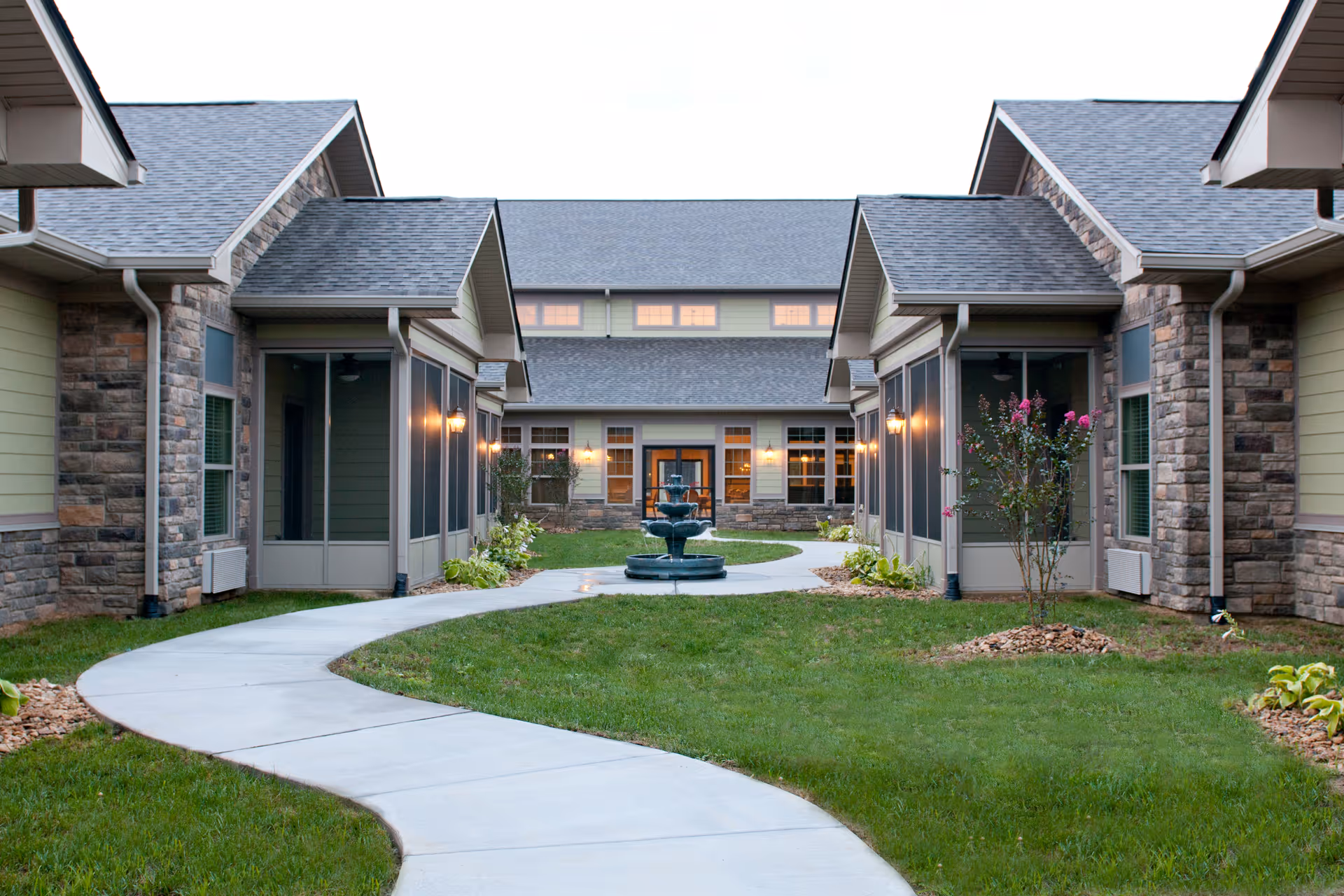 Curved concrete pathway leading through a grassy courtyard with small landscaped areas and a central water fountain, surrounded by single-story buildings with stone and siding exteriors and screened porch areas, under a cloudy sky.