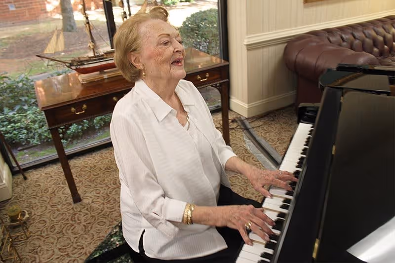 An elderly woman with short blonde hair wearing a white blouse is playing a black grand piano in a room with patterned carpet and a large window showing greenery outside. Behind her is a wooden table with a model ship on it and a brown leather tufted sofa.