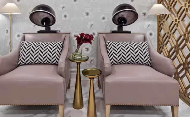 Two mauve armchairs with black and white chevron patterned pillows, each positioned under a vintage hair dryer hood in a salon-like setting. Between the chairs are two gold pedestal tables, one holding a vase with red flowers. The background features floral wallpaper and a decorative gold geometric room divider on the right.