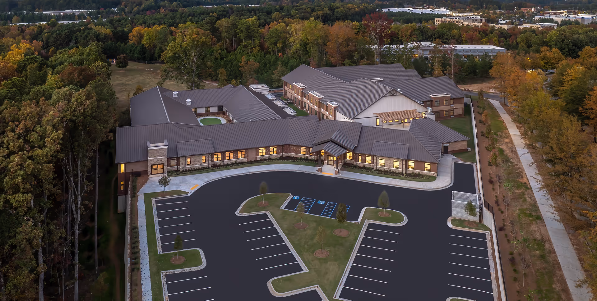 Aerial view of Mesun Senior Living facility surrounded by trees with autumn foliage. The building has a large parking lot with marked spaces, including handicapped spots, and a landscaped island with small trees. The facility is a single-story structure with a combination of brick and siding exterior and a metal roof. A paved walkway runs alongside the property.