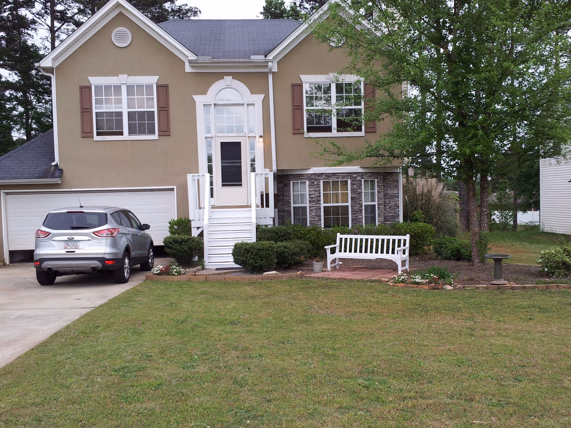 Front exterior view of a two-story beige house with white trim and brown shutters. There is a white staircase leading to the front door, a silver SUV parked in the driveway, a white bench on a small brick patio, a birdbath, and green lawn with trees and bushes.