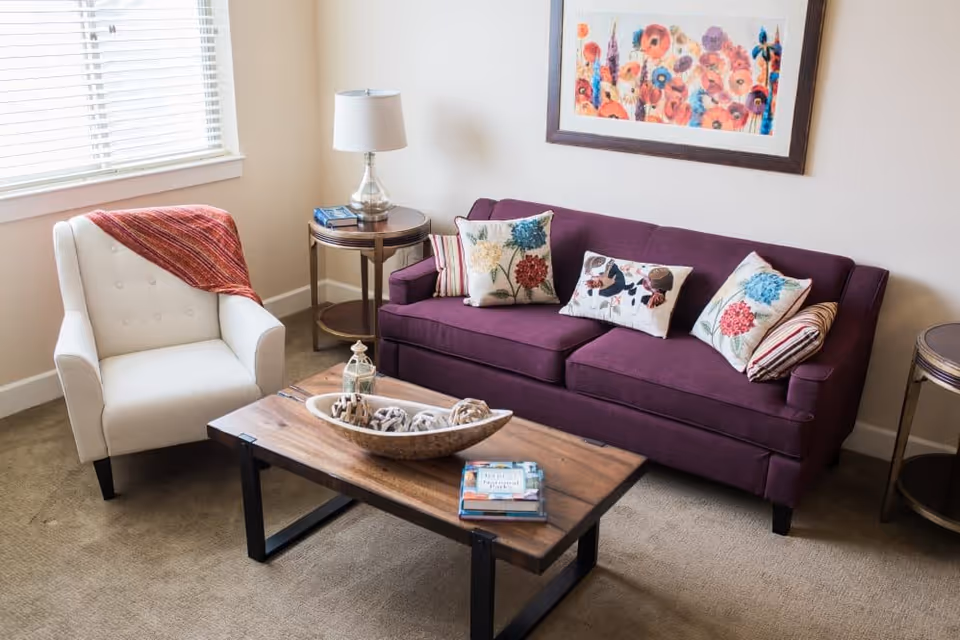 Cozy living room with a purple sofa, patterned throw pillows, a white armchair, wooden coffee table, and colorful wall art.