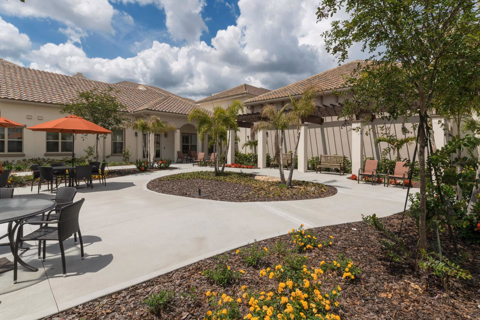 Outdoor courtyard area at The Glades at ChampionsGate featuring a circular concrete walkway with a central garden bed containing small palm trees. Surrounding the walkway are patio tables with chairs and orange umbrellas, benches under a pergola, and landscaped flower beds with yellow flowers. The building has a tiled roof and beige exterior walls under a partly cloudy sky.