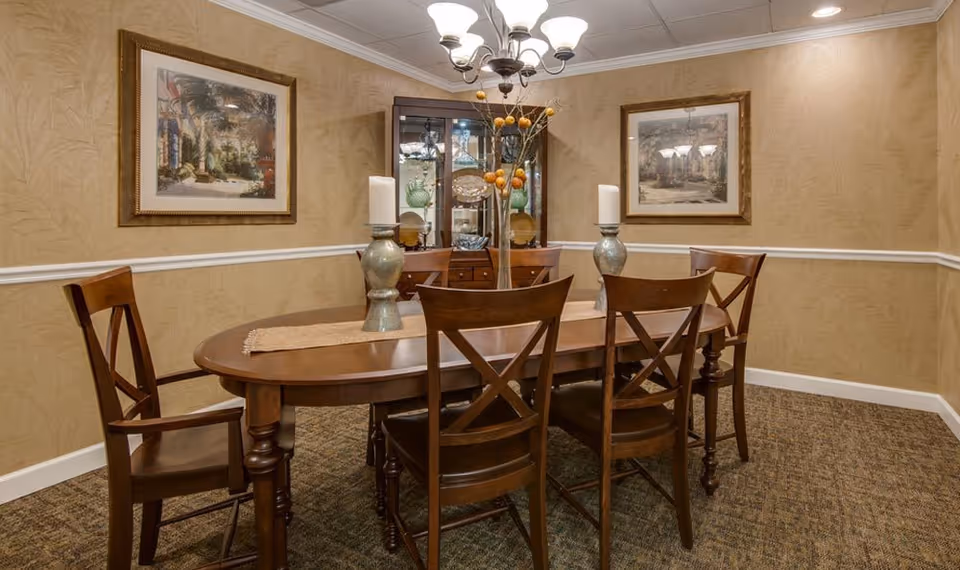 A dining room with a wooden oval table surrounded by six wooden chairs. The table has a beige runner with two large candle holders and a tall vase with decorative branches. The walls are beige with a white chair rail, and two framed paintings hang on the walls. A wooden china cabinet with glass doors is positioned against the back wall, displaying glassware and dishes. A chandelier with multiple lights hangs from the ceiling.