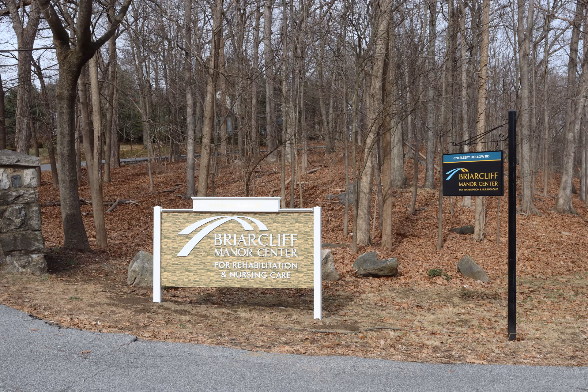 Outdoor view of two signs for Briarcliff Manor Center for Rehabilitation and Nursing Care located near a wooded area with leafless trees and dry leaves on the ground. One sign is large and mounted on white posts, the other is smaller and hanging from a black metal post.