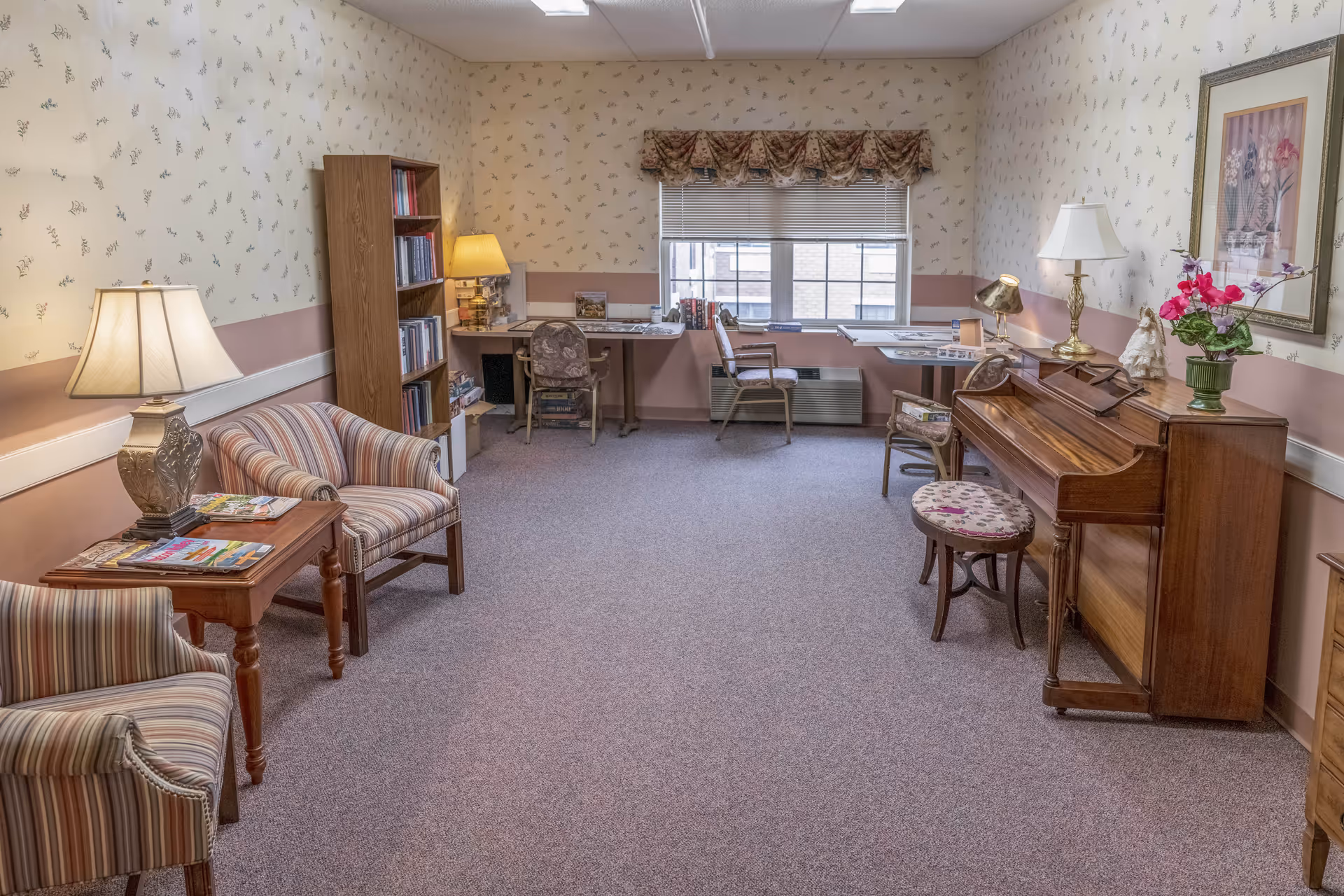 A cozy room with floral wallpaper and carpeted floor featuring two striped armchairs and a wooden side table with a lamp and magazines. There is a wooden bookshelf filled with books, two desks with chairs near a window with blinds and a floral valance, and a wooden piano with a floral cushioned stool. The room is softly lit and decorated with a framed floral picture and a vase of flowers on the piano.