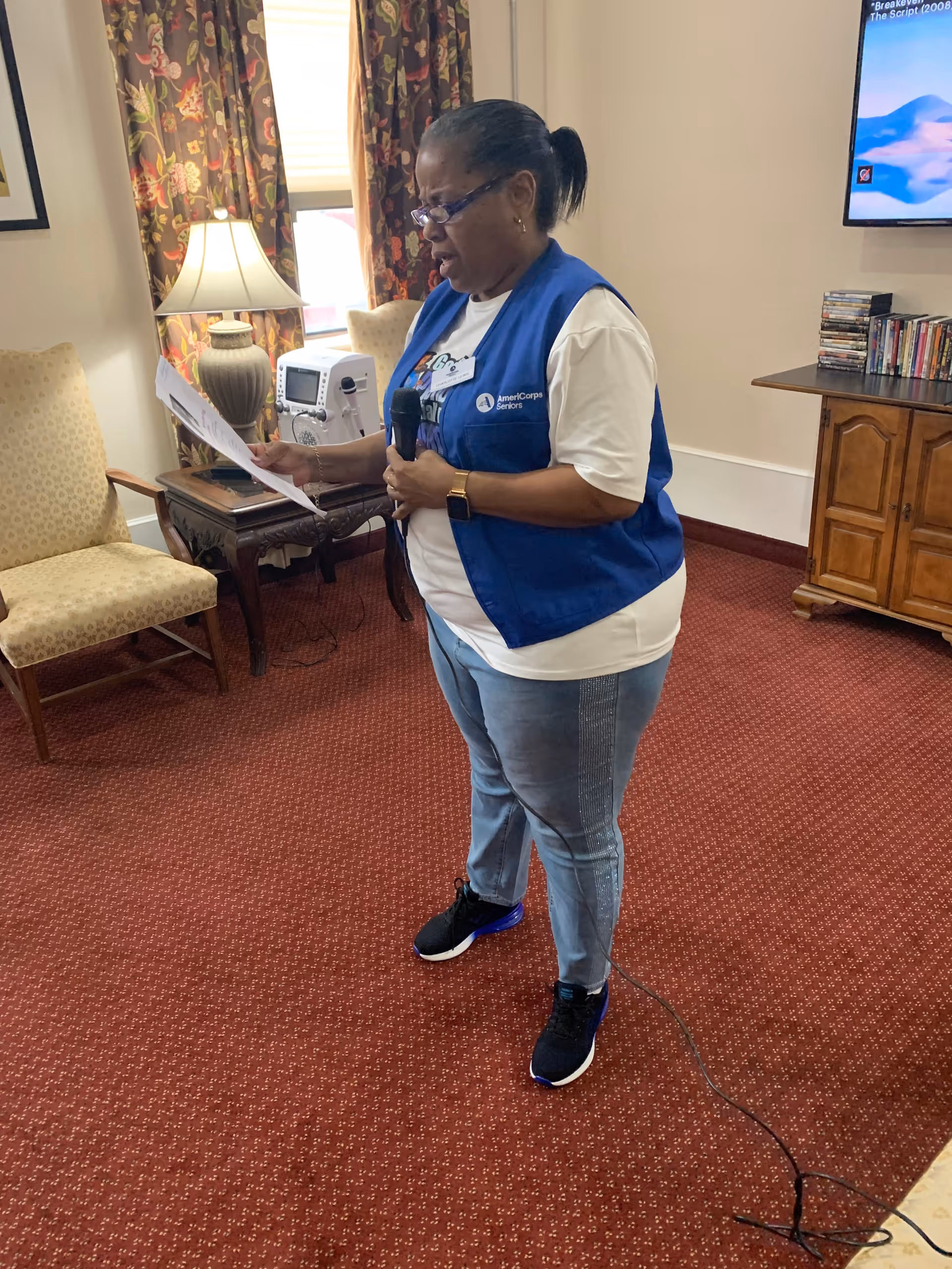 A woman wearing a blue AmeriCorps Seniors vest and glasses is standing on a red carpeted floor inside a room. She is holding a microphone in one hand and reading from a sheet of paper in the other. Behind her, there is a beige upholstered chair, a wooden side table with a lamp and medical equipment, floral curtains covering a window, and a wooden cabinet with DVDs and a TV mounted on the wall.