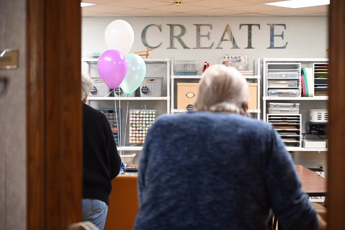 Two elderly individuals seen from behind in a room with shelves filled with craft supplies and the word 'CREATE' displayed on the wall. There are three balloons in white, purple, and light green colors near the shelves.