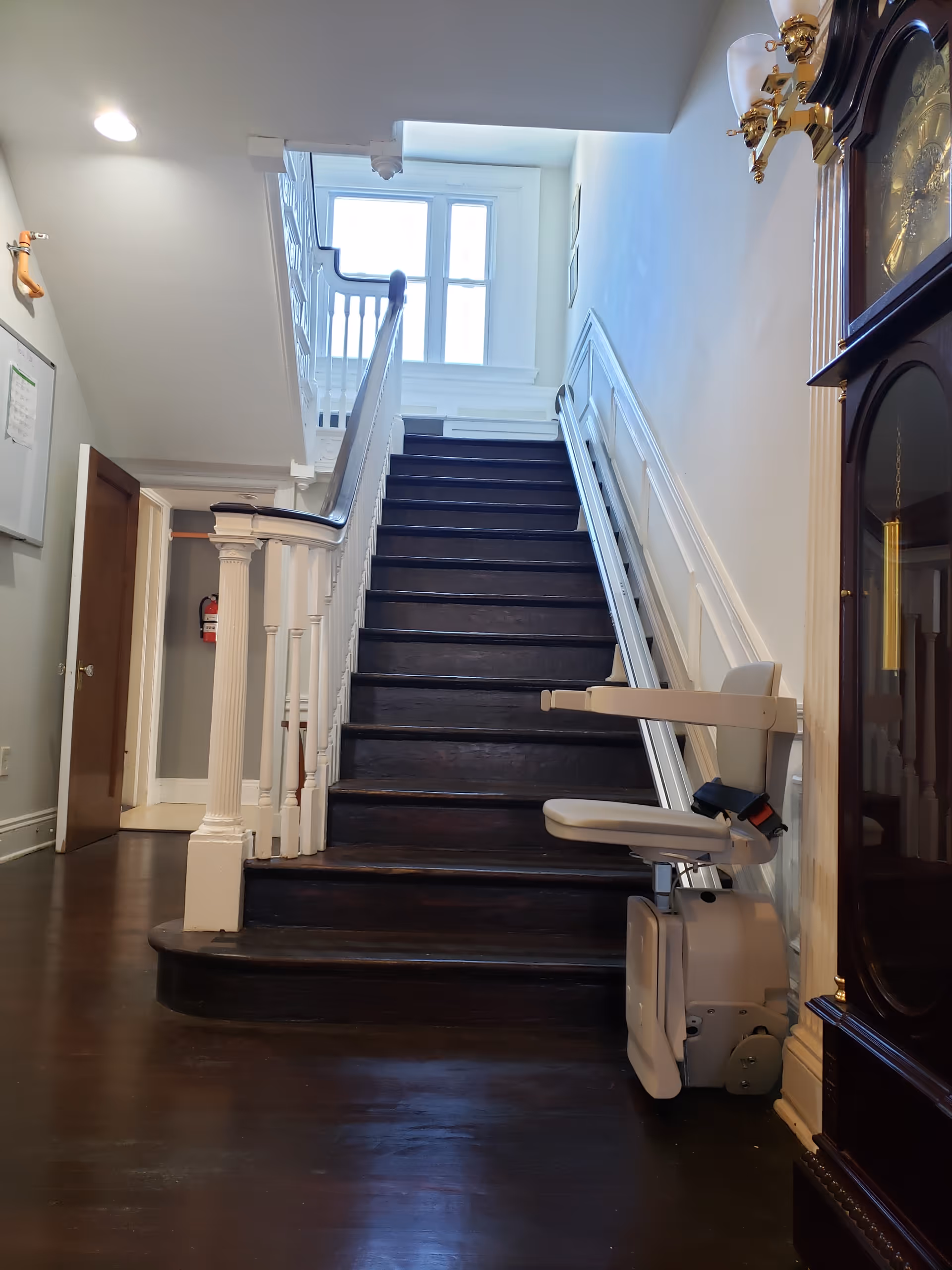 Interior view of a stairway with dark wooden steps and white railings. A stair lift chair is installed on the right side of the stairs. There is a large window at the top of the stairs letting in natural light. To the right of the stairs is a tall wooden grandfather clock. The floor is dark wood, and there is a door open to a room on the left side of the image.