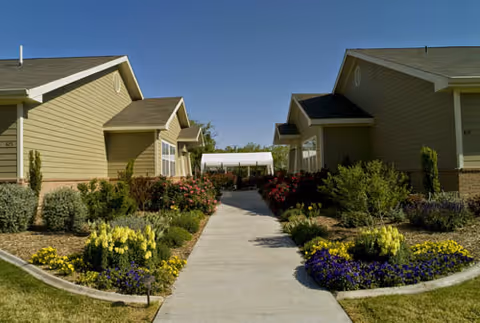 Concrete walkway between two single-story beige buildings with landscaped flower beds leading to a covered entrance.