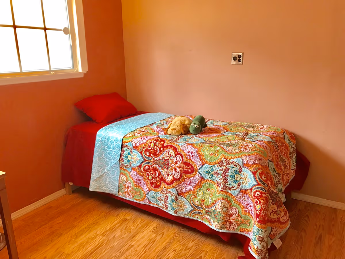 A small bedroom with a single bed covered in a colorful patterned quilt and a light blue blanket. There are two stuffed toys placed on the bed. The room has peach-colored walls, a window with white trim letting in natural light, and a wooden floor.