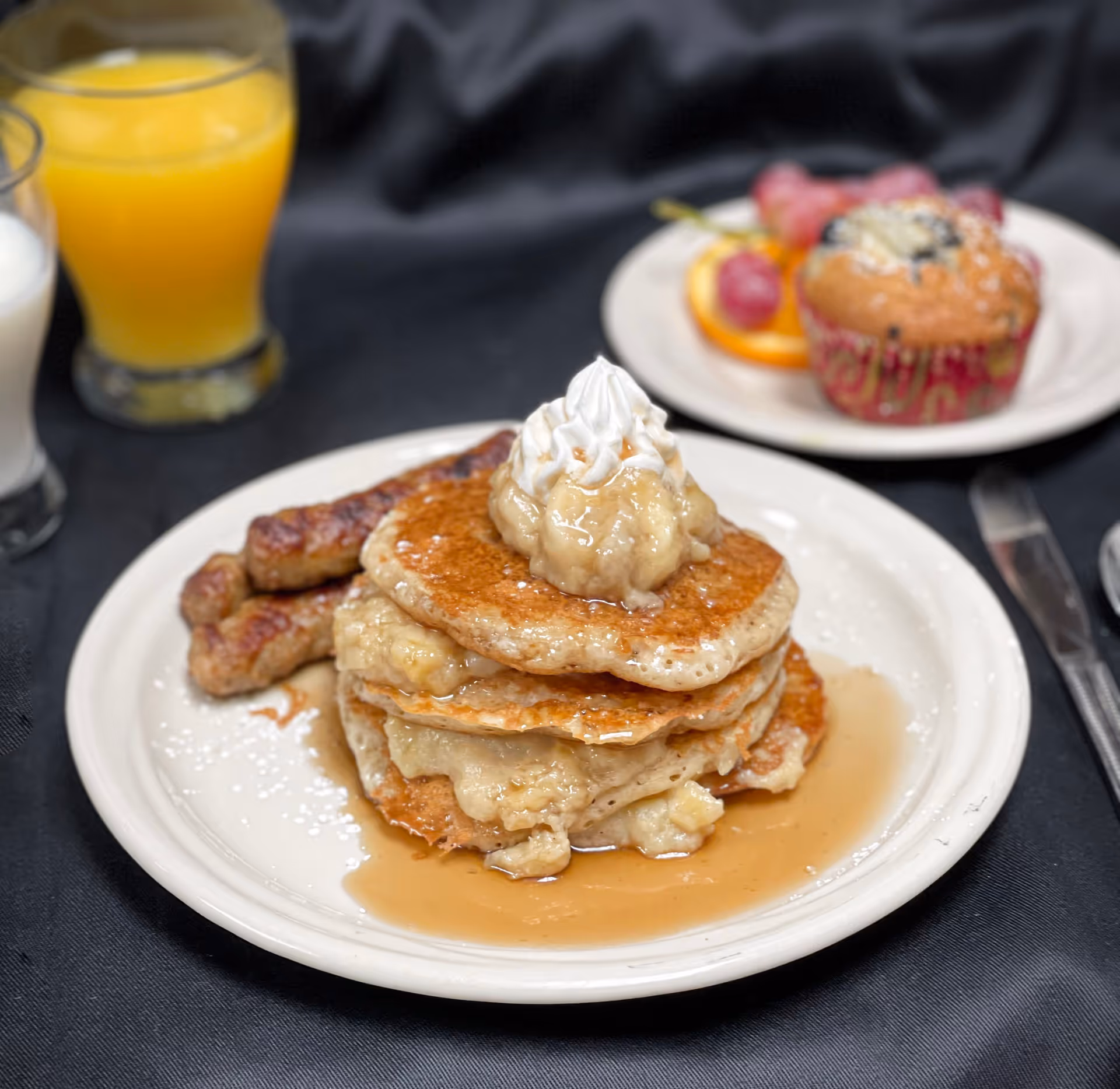 A stack of pancakes topped with whipped cream and banana compote on a plate with sausages, plus glasses of orange juice and milk and a muffin in the background.