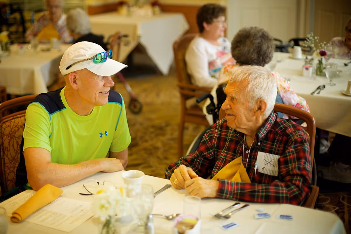 Two men, one elderly and one wearing a bright green shirt and white cap, sit and converse at a set dining table in a retirement community dining room.