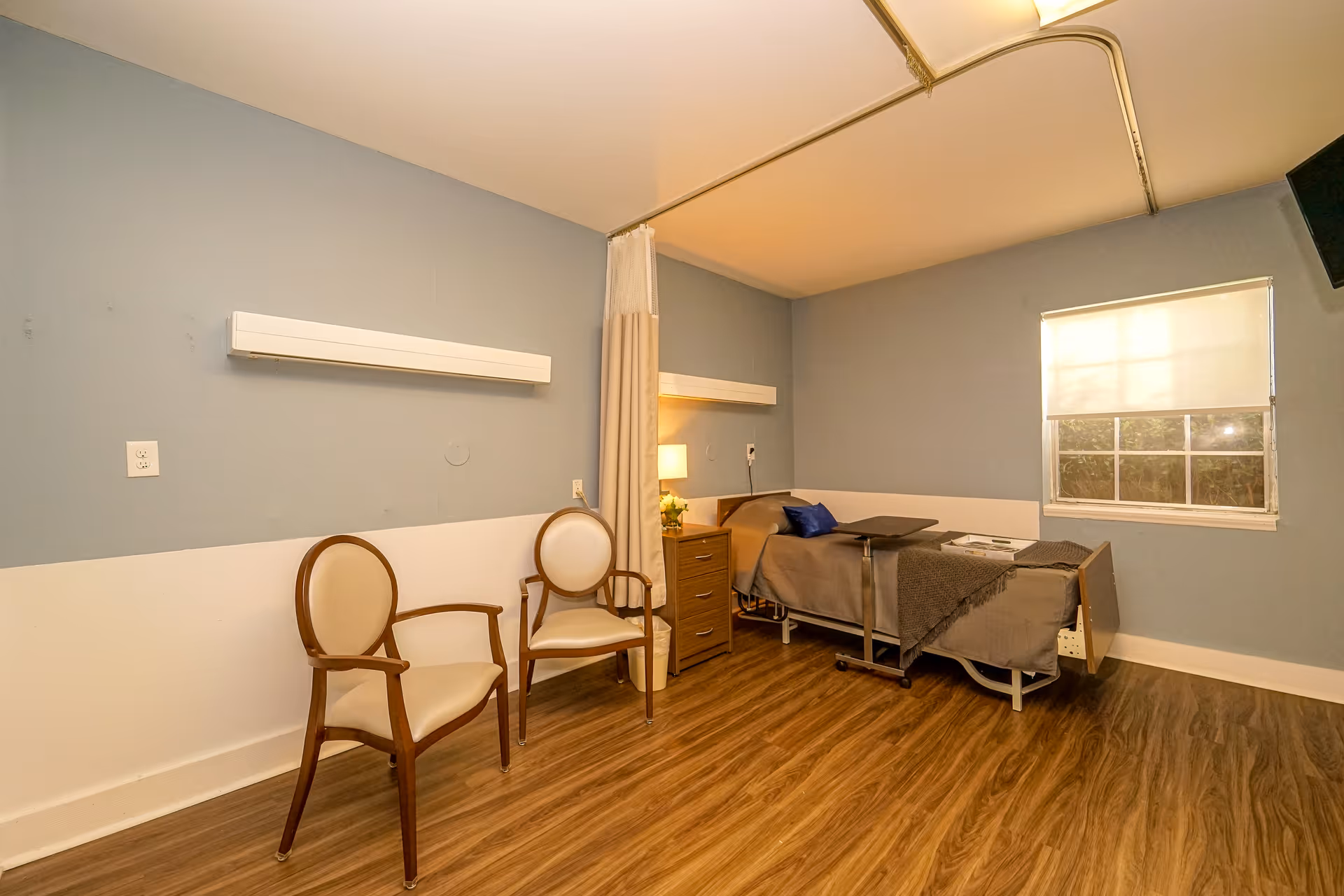 A nursing facility room with two wooden chairs with cream cushions, a hospital bed with a gray blanket and a blue pillow, a bedside table with a lamp and flowers, a window with a white blind, and wood-patterned flooring. The walls are painted blue and white, and there is a ceiling-mounted curtain rail for privacy.
