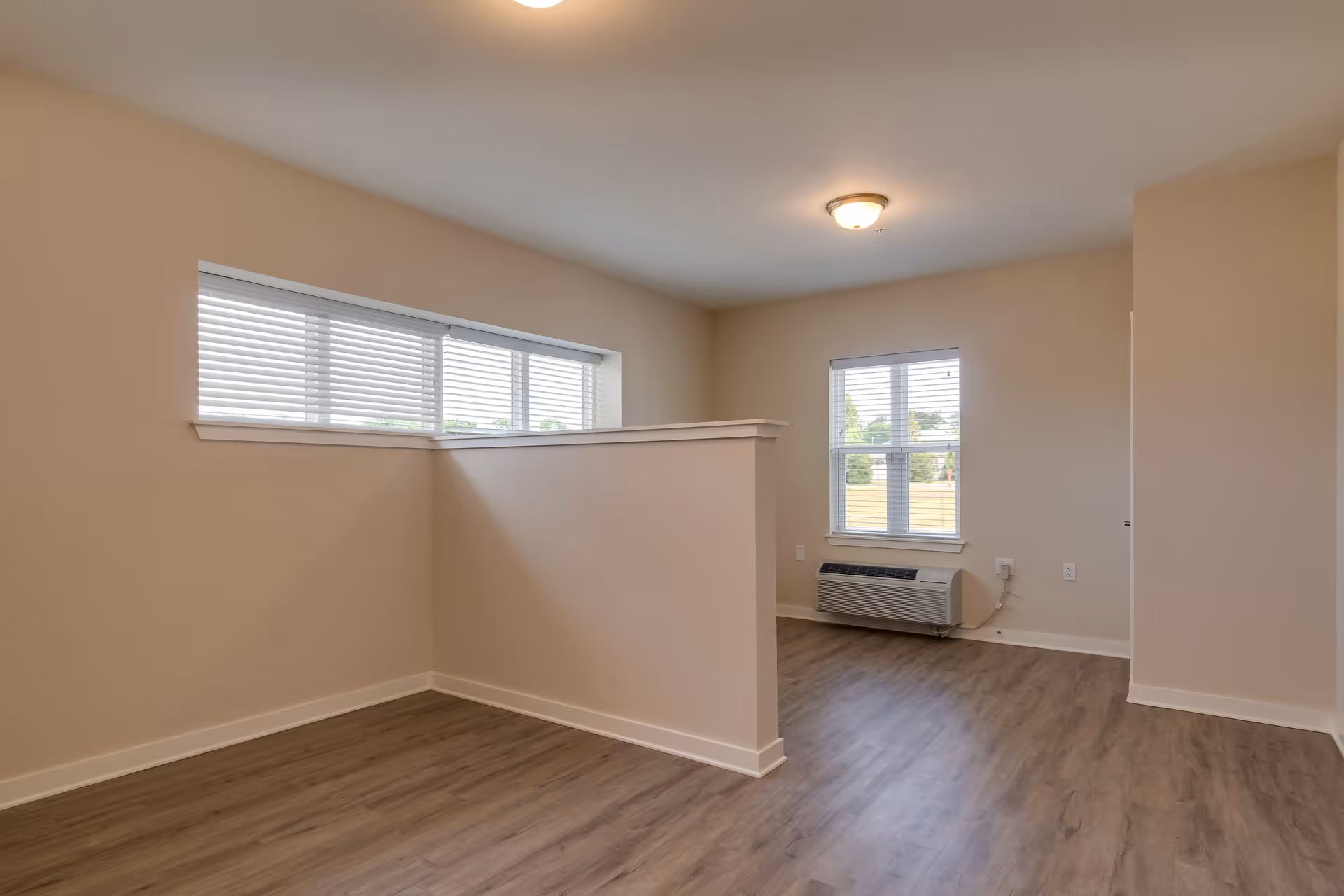 Empty interior room with light beige walls, wood flooring, a half wall partition, two windows with white blinds, and a wall-mounted air conditioning unit below one window.