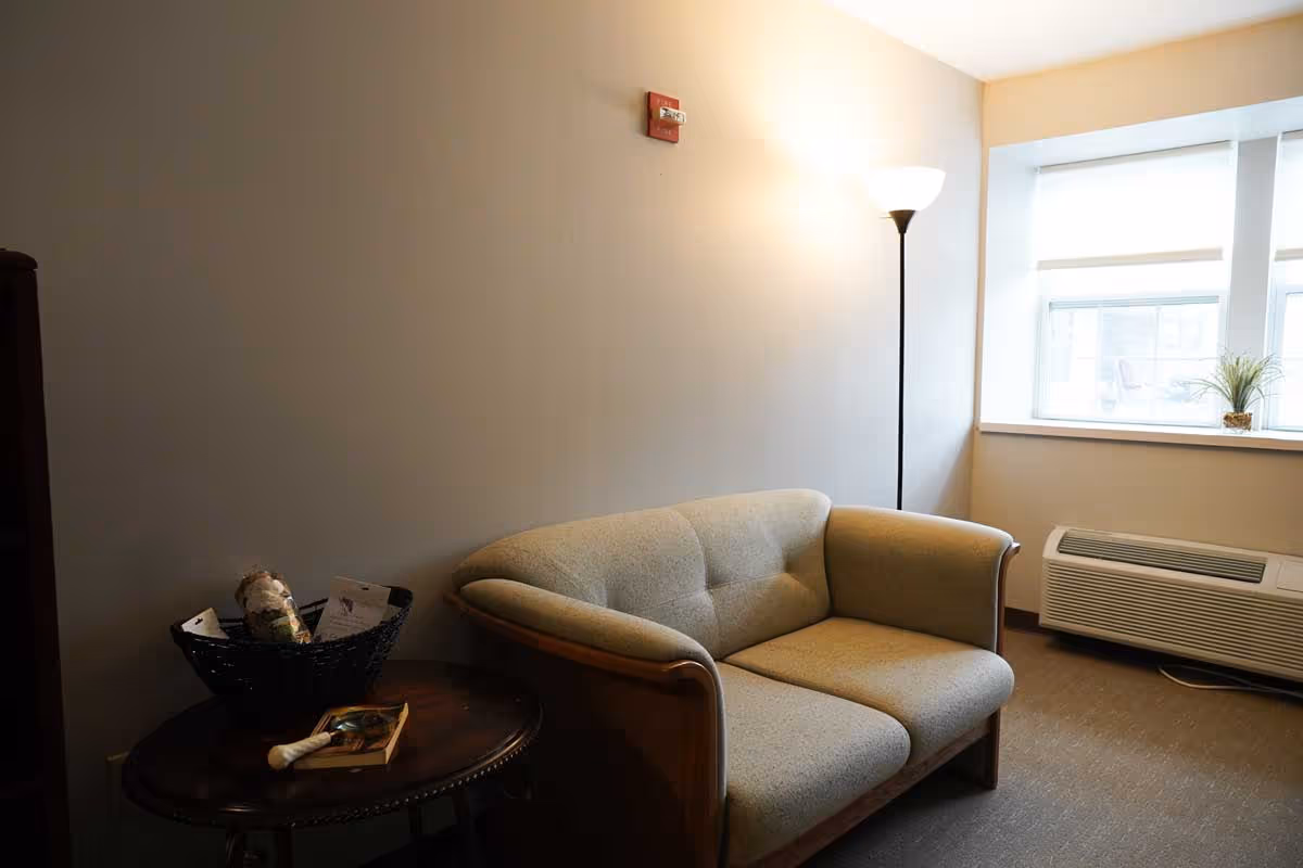 A small sitting area with a beige loveseat, a round wooden side table holding a basket and a book, a tall floor lamp, and a window with blinds letting in natural light. There is an air conditioning unit below the window.