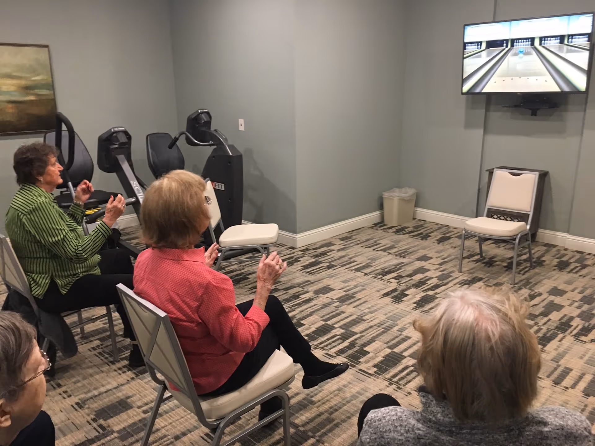 A group of elderly women seated on chairs in a room watching a screen mounted on the wall displaying a virtual bowling lane. The room has exercise equipment against one wall and a patterned carpeted floor.