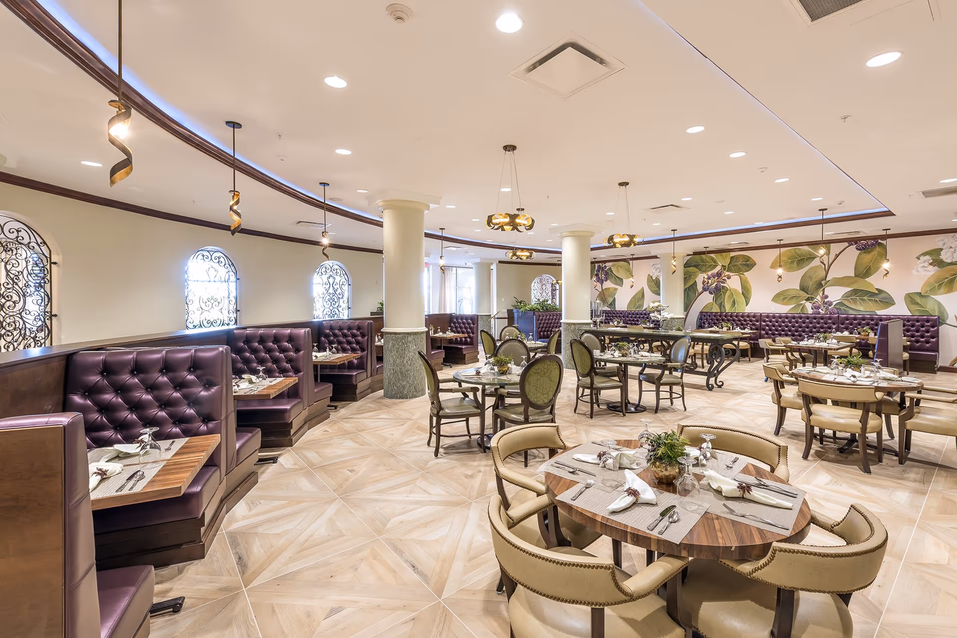 Spacious and elegant dining room with round and booth seating. The room features tufted purple leather booths along the wall, round wooden tables with beige and green upholstered chairs, and decorative hanging light fixtures. The walls have large botanical murals with green leaves and purple berries, and the floor is tiled with a light wood pattern.