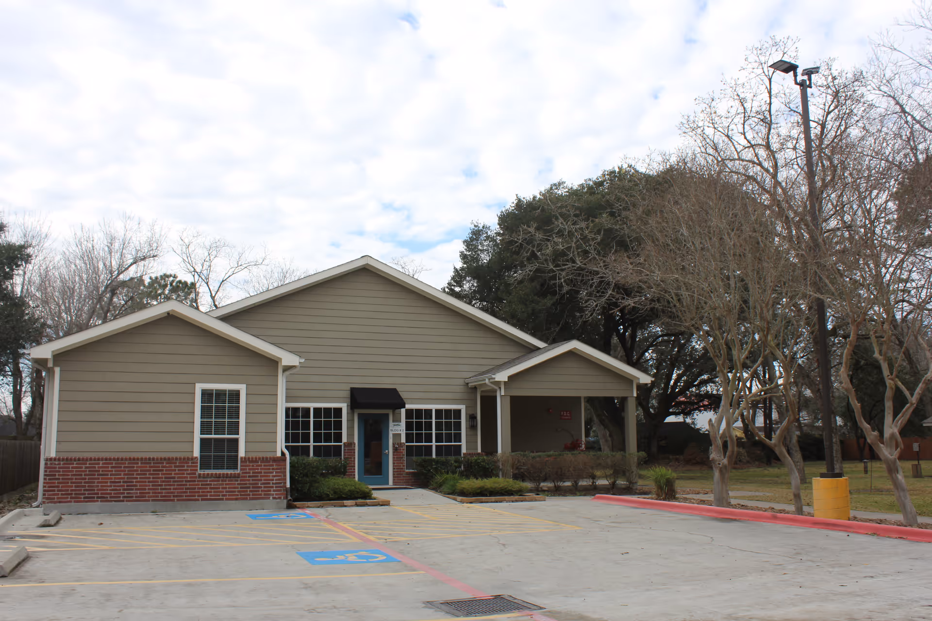 Exterior view of a single-story building with beige siding and red brick accents at the base. The building has a blue door with a black awning above it, several windows, and a covered entrance area. There are parking spaces in front, including two handicap parking spots marked in blue. Leafless trees and a tall street lamp are visible around the building under a cloudy sky.