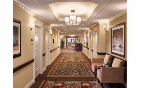 A well-lit, carpeted hallway in a senior living facility with beige walls, white doors, framed artwork, wall sconces, and a chandelier. There are two cushioned chairs with a small table holding a bowl of fruit and a flower arrangement on the right side. The hallway extends into the distance with more plants and furniture visible at the far end.
