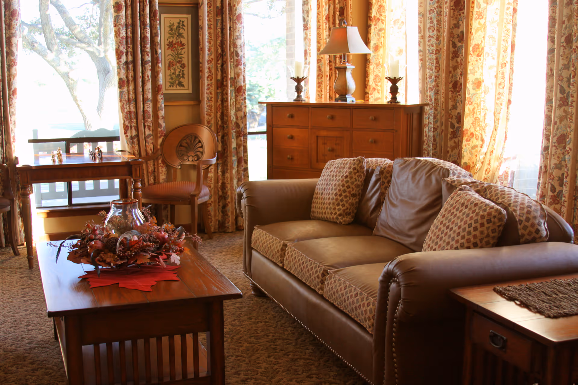 A cozy living room with a brown leather sofa adorned with patterned cushions, a wooden coffee table decorated with a floral centerpiece, a wooden side table with a woven mat, a wooden chair, and a wooden dresser with candles and a lamp. The room is warmly lit by natural light coming through large windows with floral curtains.