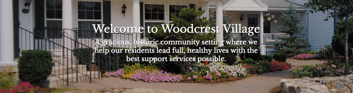 Front exterior view of Woodcrest Village Assisted Living showing a welcoming entrance with white columns, a ramp with railings, bushes, and colorful flower beds along the pathway.