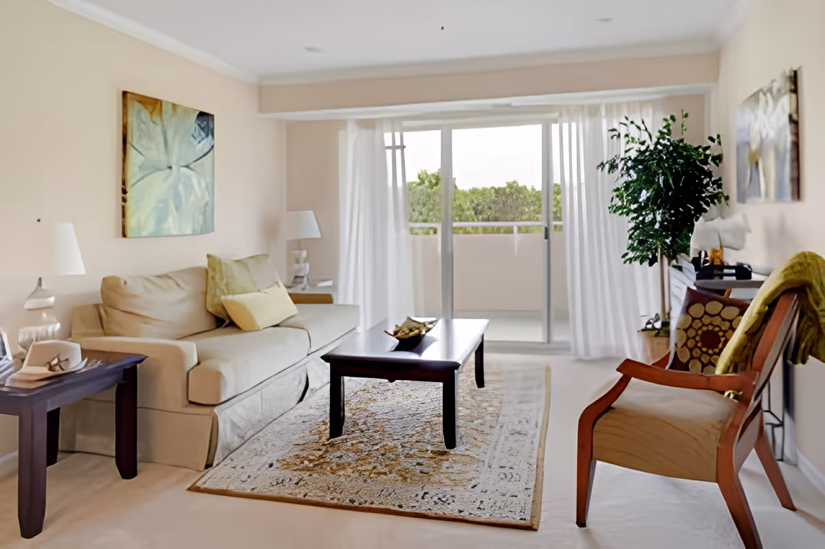 A bright living room with a beige sofa adorned with cushions, a wooden coffee table on a patterned rug, a wooden armchair with a green throw, two table lamps on side tables, a large potted plant, and sliding glass doors with sheer white curtains leading to a balcony with greenery outside.