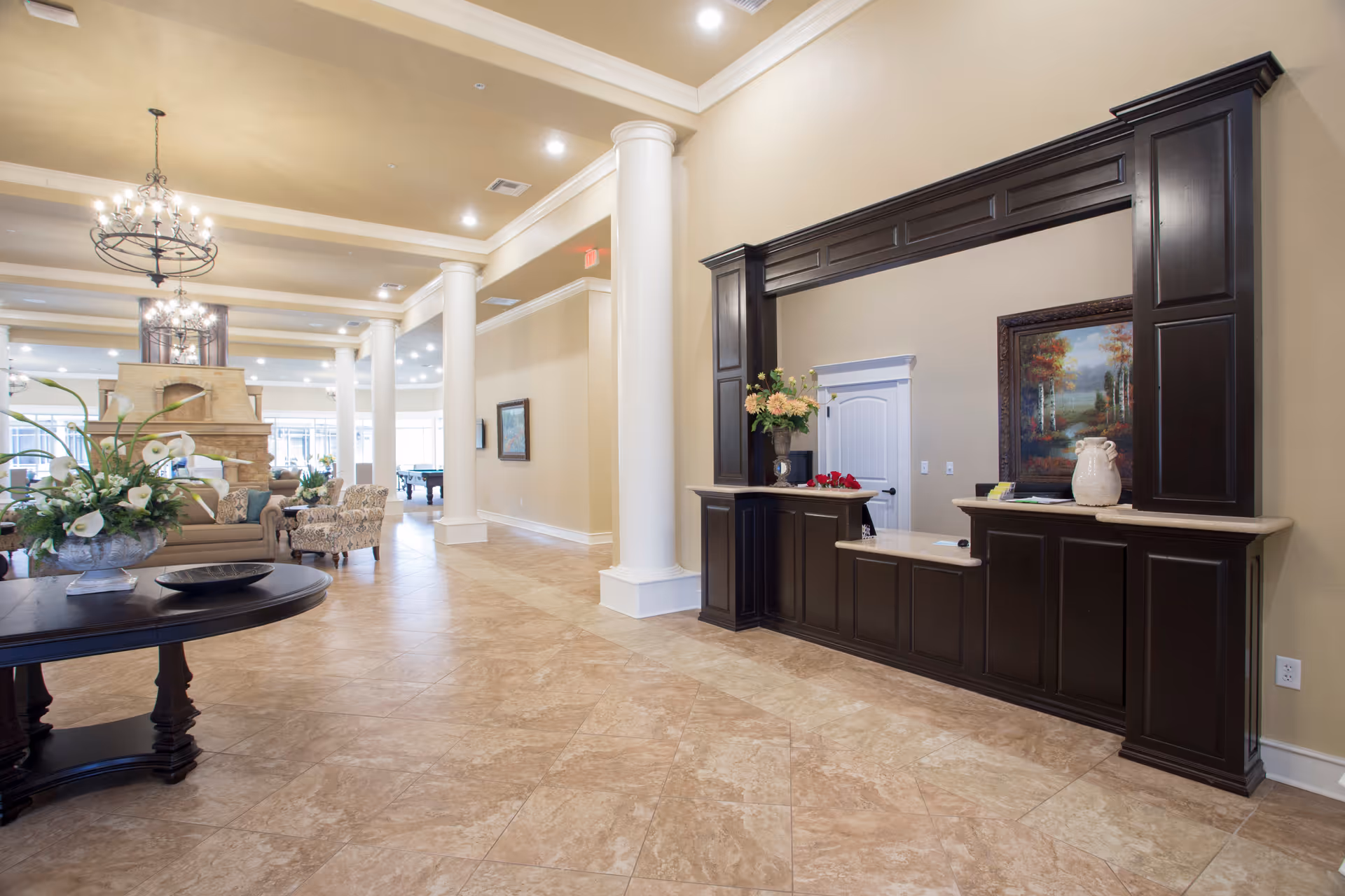 Spacious and elegant senior living facility lobby with beige tiled floors, tall white columns, a dark wood reception desk with decorative vases and flowers, a large painting on the wall, comfortable seating area with sofas and armchairs, and ornate chandeliers hanging from the ceiling.