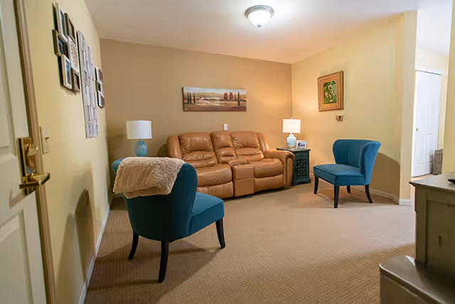 Cozy living room with a tan leather sofa, two blue accent chairs, side tables with lamps, and wall art on beige walls.