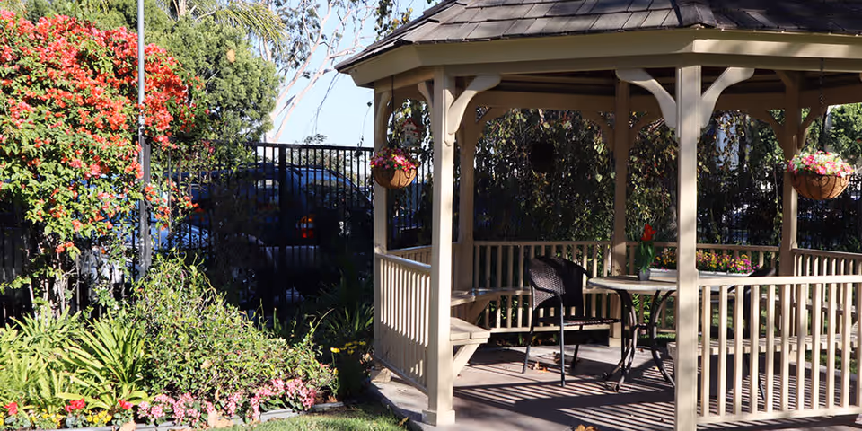 A beige wooden gazebo in a garden area with hanging flower baskets and a table with chairs inside. Surrounding the gazebo are various green plants and colorful flowers, with a black metal fence and trees in the background.
