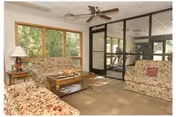 Sunny common room with floral sofas, a coffee table, large windows, and a glass-walled exercise room beyond.