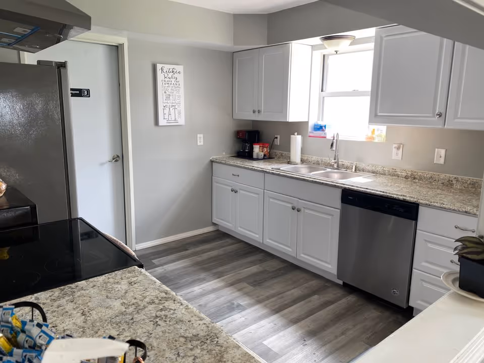 A modern kitchen with white cabinets, granite countertops, a double sink under a window, a stainless steel dishwasher, and a black electric stove. There is a coffee maker and some kitchen items on the counter, a door labeled 'Employees' in the background, and a decorative sign on the wall.