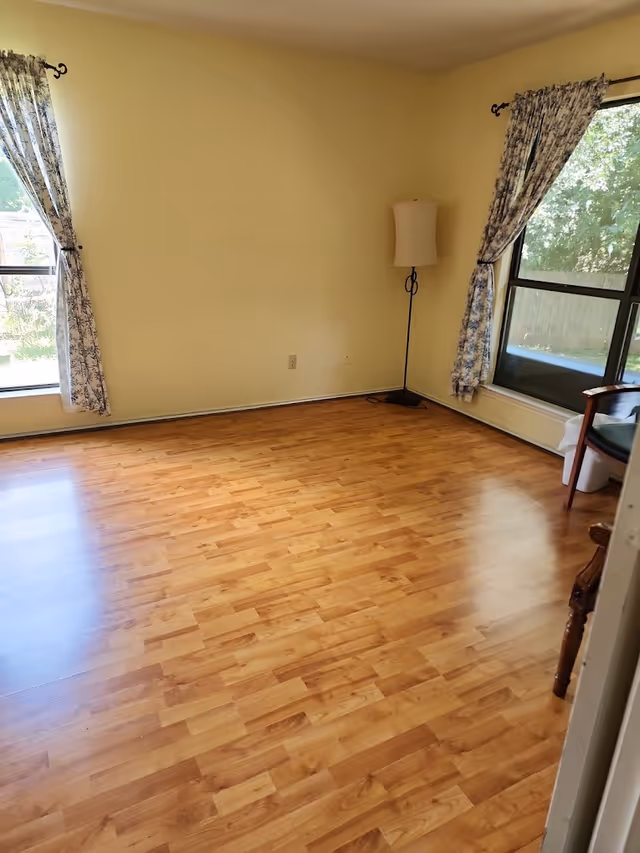 Empty room with wood laminate flooring, two large windows with floral curtains, and a floor lamp near the corner.