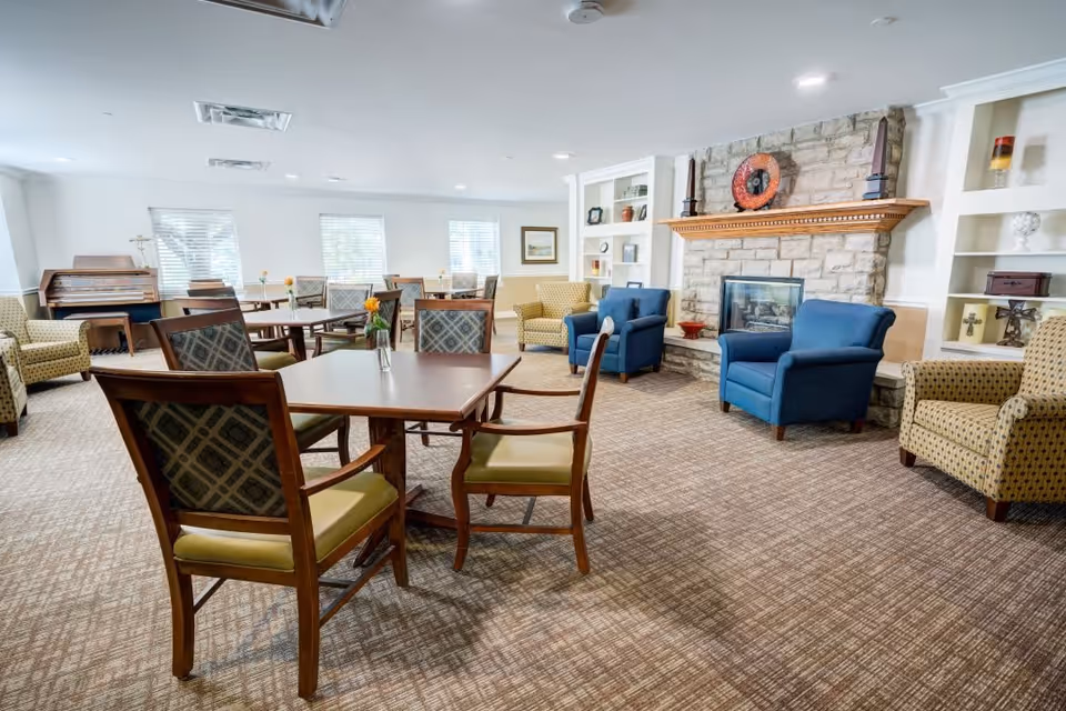 A spacious, well-lit common area with multiple wooden tables and cushioned chairs arranged neatly. The room features a stone fireplace with a wooden mantle, flanked by built-in shelves displaying decorative items. There are several comfortable armchairs in blue and patterned upholstery around the fireplace. Large windows allow natural light to fill the room, and a piano is visible in the background.