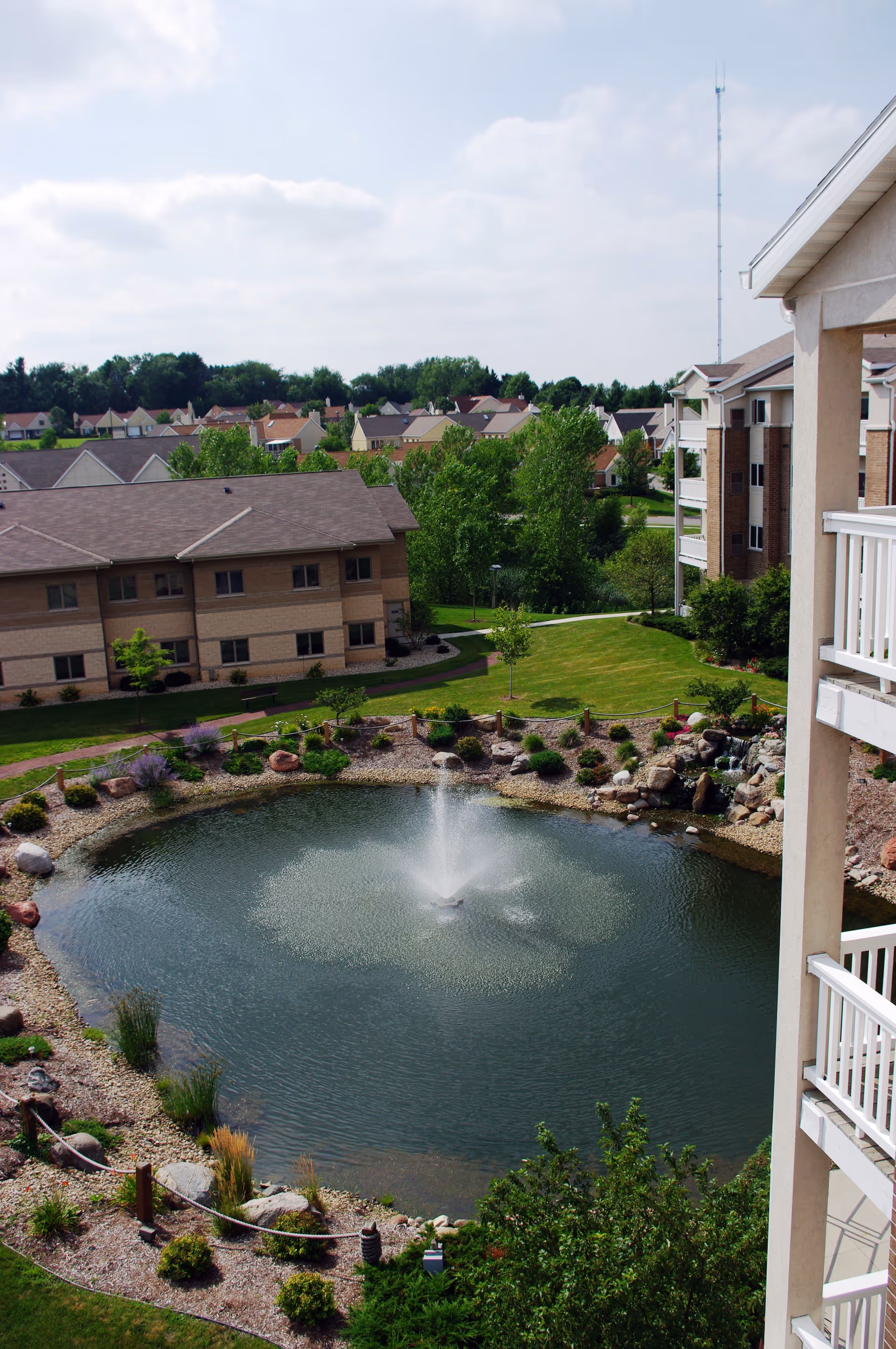 View of a landscaped pond with a central water fountain surrounded by rocks, plants, and a walking path. Residential buildings and trees are visible in the background under a partly cloudy sky.