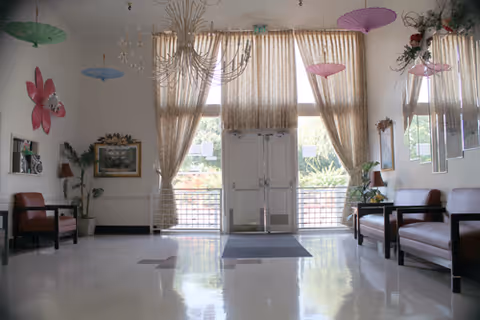 Bright, spacious lobby/entrance with seating benches, tall windows draped with curtains, chandeliers, and decorative hanging parasols.
