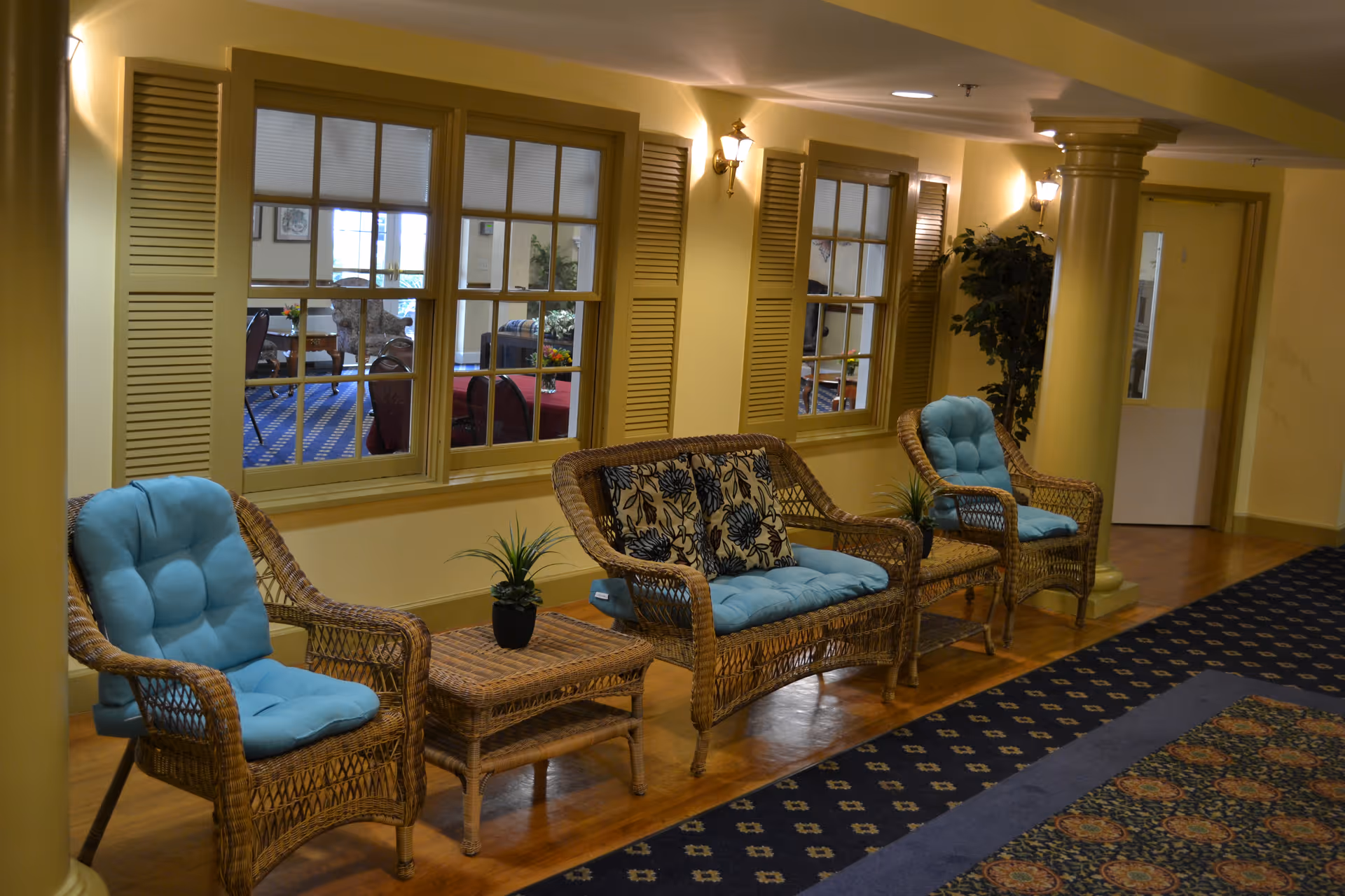 Wicker seating area with blue cushions and small tables in a warmly lit lounge corridor with windows and decorative columns.