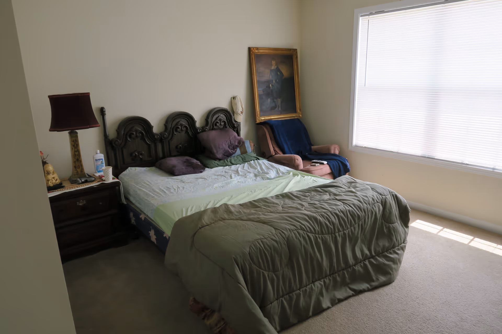 A bedroom with a large bed featuring an ornate dark wooden headboard, several pillows, and a green comforter. Next to the bed is a dark wooden nightstand with a lamp, a bottle of lotion, and other small items. In the corner, there is a pink armchair draped with a blue blanket and a framed painting leaning against the wall. A large window with closed blinds allows natural light to fill the room.