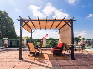 Outdoor patio area with a pergola providing shade over a seating arrangement including chairs and a small table. The patio is paved with large tiles and has a railing with potted plants. An American flag is visible in the background under a partly cloudy blue sky.