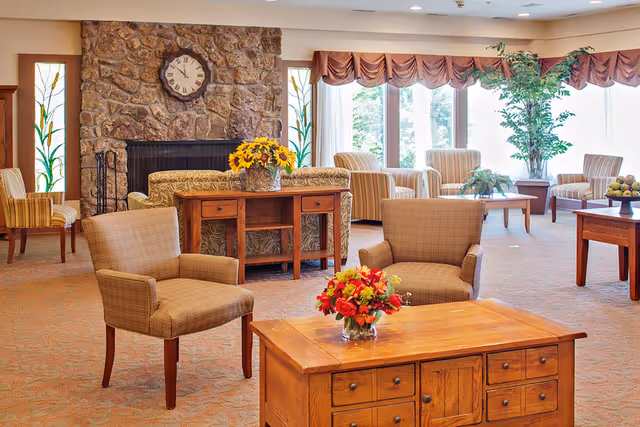Bright senior living facility sitting area with armchairs, wooden tables, a stone fireplace and large windows.