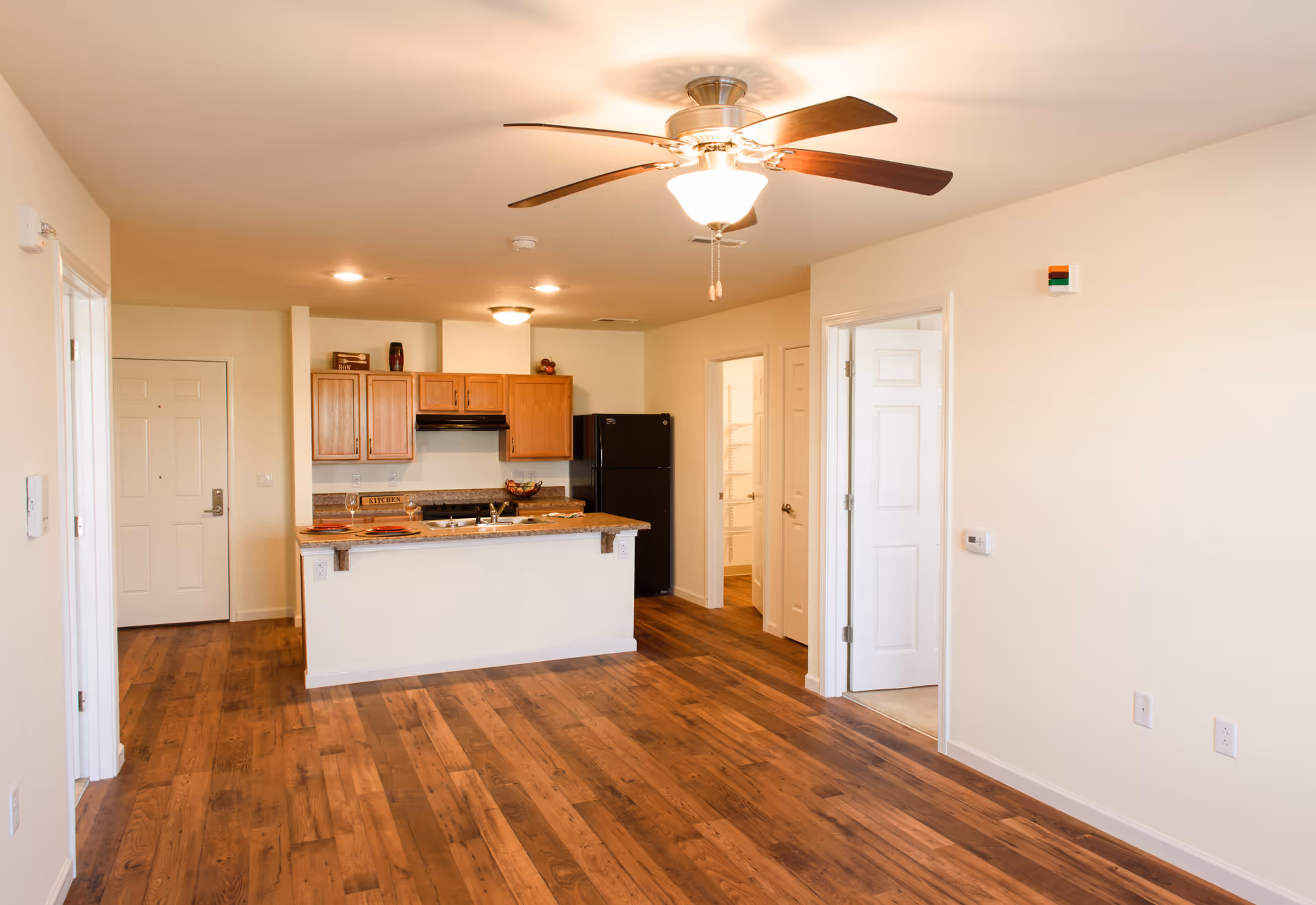 Open-plan apartment interior with hardwood floors, a ceiling fan, and a kitchen area featuring a breakfast island, wooden cabinets, and a black refrigerator.