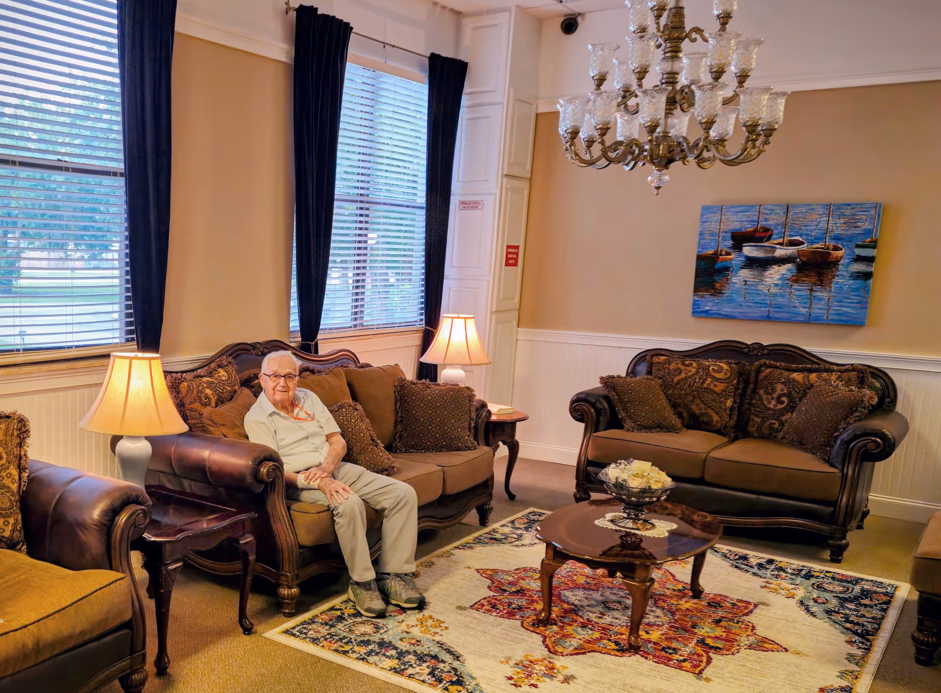 An older adult sits on a sofa in a warmly decorated communal living room with lamps, a chandelier, sofas and a colorful rug.