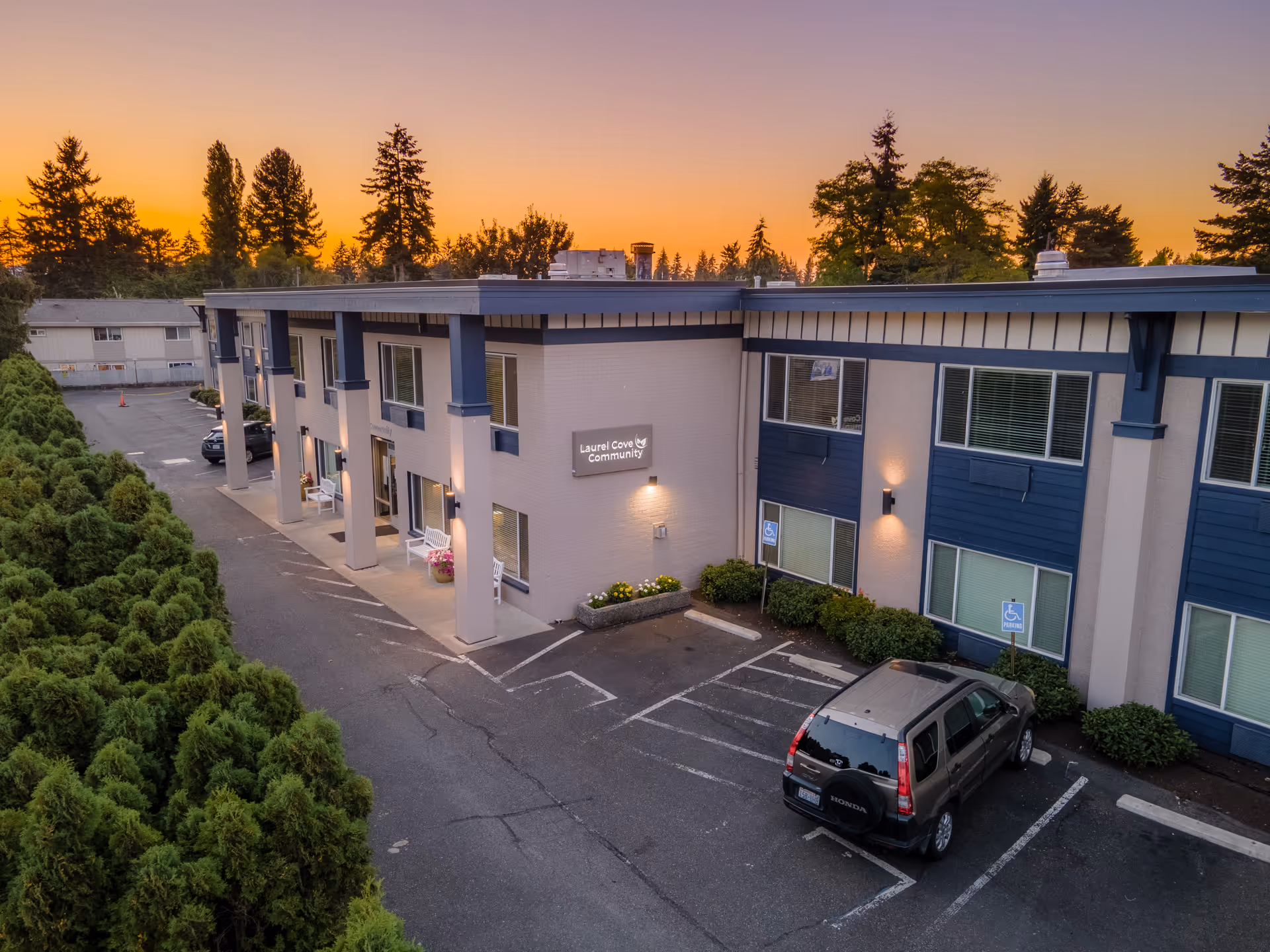Exterior view of Laurel Cove Community assisted living and senior care building at sunset, showing a two-story structure with parking spaces, including handicapped spots, and surrounded by trees and greenery.