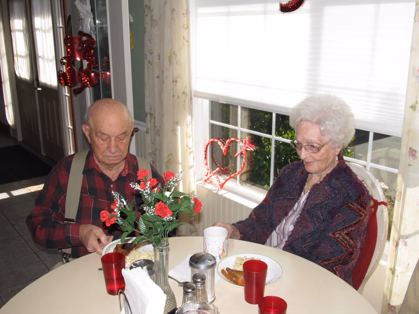 An elderly man and woman sitting at a round dining table with plates of food and red cups. The man is wearing a red and black plaid shirt with suspenders, and the woman is wearing glasses and a patterned shawl. There is a vase with red flowers on the table, and a window with floral curtains and a heart-shaped decoration in the background.