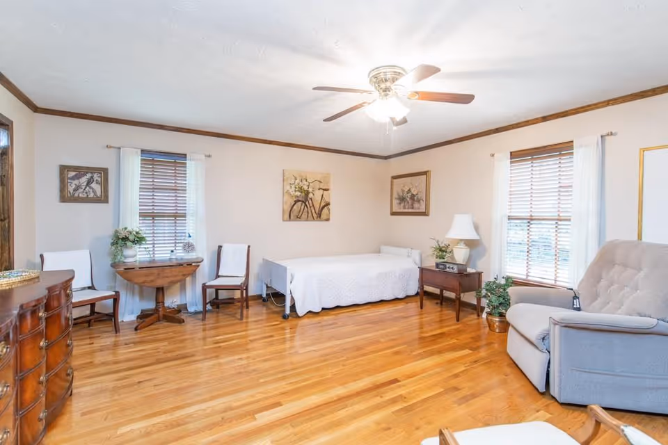 A bright and spacious room with hardwood floors featuring a single bed with a white quilt, two windows with wooden blinds and white curtains, a wooden dresser, a small round wooden table with two chairs, a side table with a lamp and plant, and a comfortable gray recliner chair. The walls are decorated with framed artwork and the ceiling has a fan with lights.
