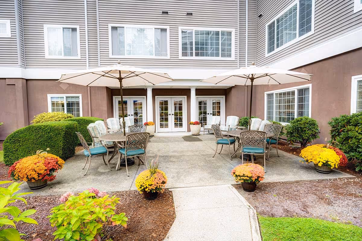 Outdoor patio area at Gig Harbor Court with tables and chairs under large umbrellas, surrounded by potted flowers and neatly trimmed bushes, adjacent to a building with multiple windows and glass doors.