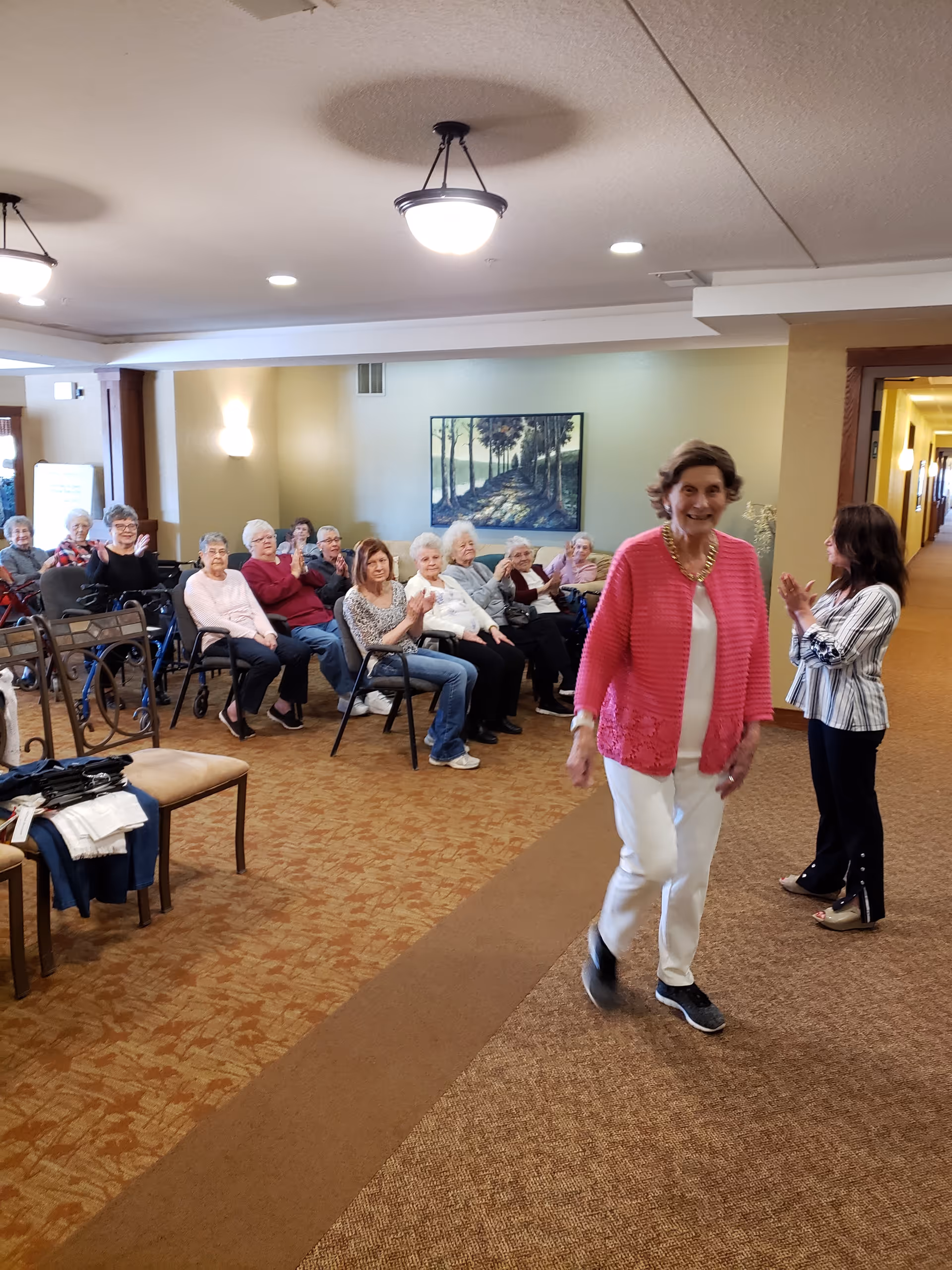 A group of elderly women seated in chairs along the wall of a well-lit room with beige walls and carpet. One woman in a pink cardigan and white pants is walking towards the camera, smiling, while another woman stands nearby clapping. A painting of a tree-lined path hangs on the wall behind the seated group.