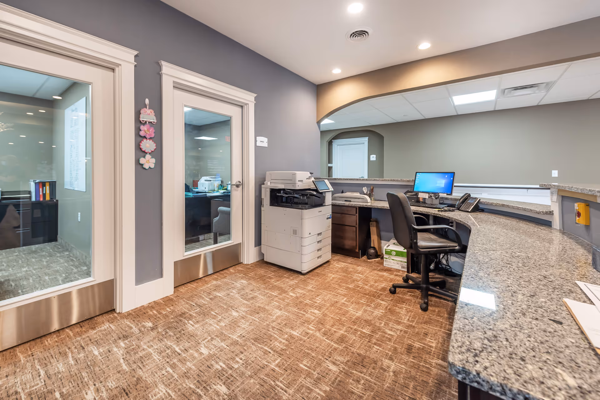 Reception area with a curved granite counter, office chair and computer, a copier, and glass office doors.